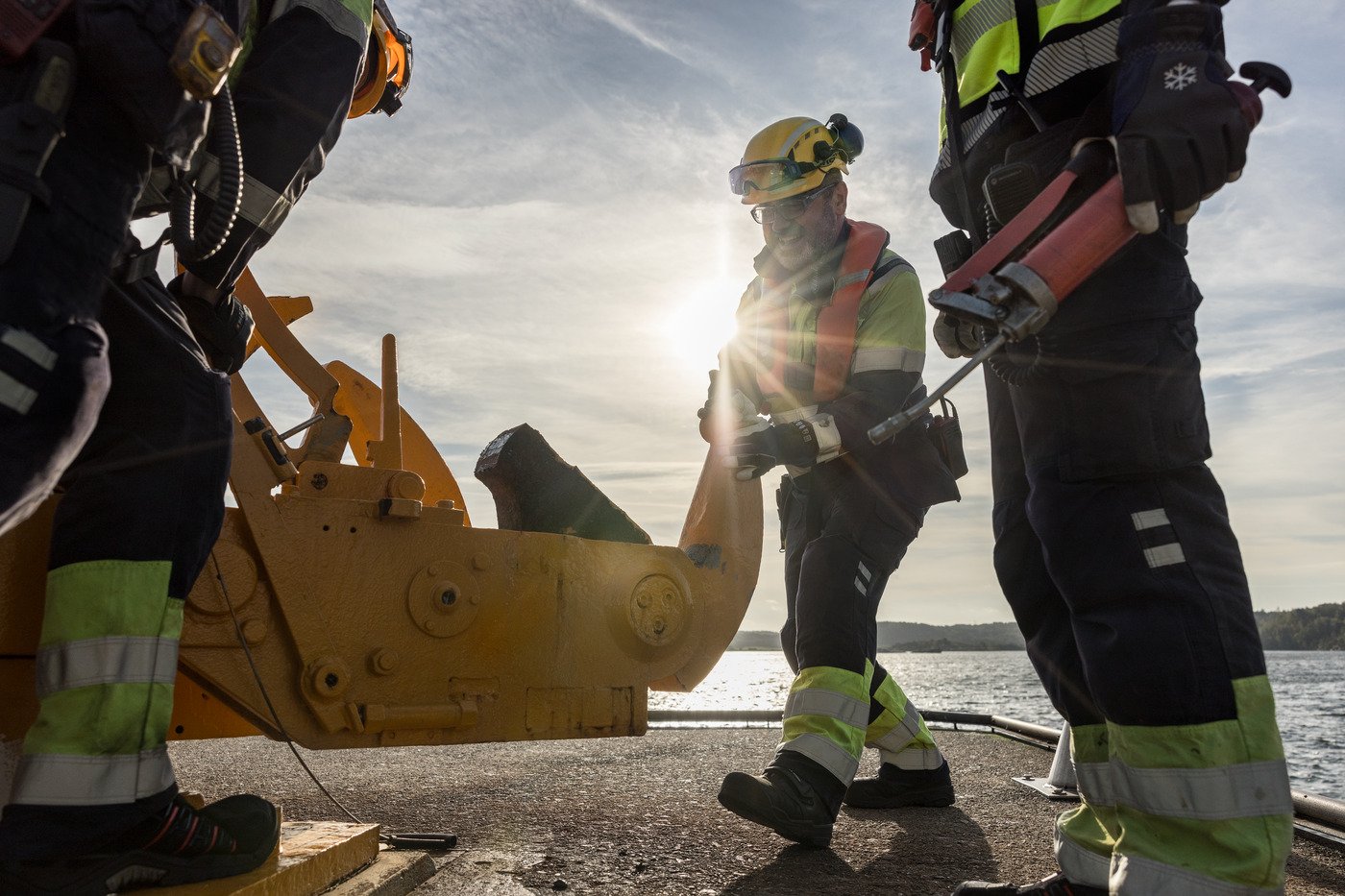 3 hamnarbetare gör underhåll på maskin i hamnen, motljus